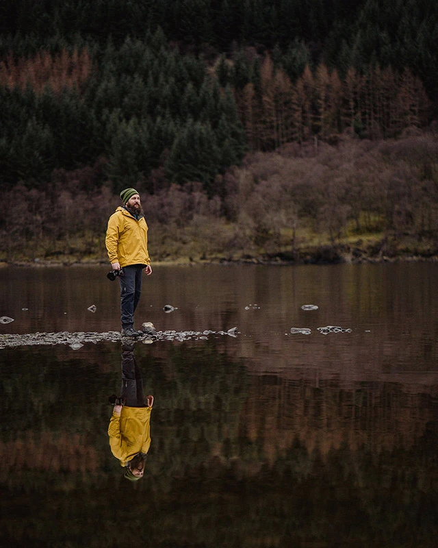 Weirdie Grizzly Scotland elopement photographer stands at the lake side surrounded be a huge body of water with pine forest in the background